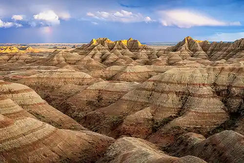 Clearing Storm and Rainbow - Badlands National Park Late afternoon photo in Badlands National Park showing layered red and white hills, storm clouds, and a faint rainbow in the distance with light touching faraway terrain. Image by Rick Russell.