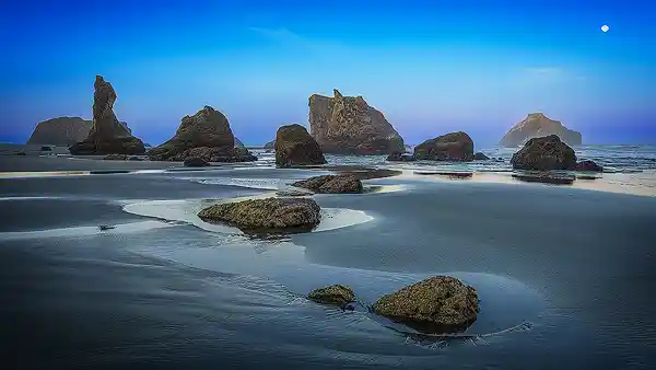 Sunrise Panorama - Bandon Beach Panoramic sunrise photo of Bandon Beach with tide pools reflecting blue and pink hues, distinct sea stacks in the distance, and the moon in a clear sky. Image by Rick Russell.