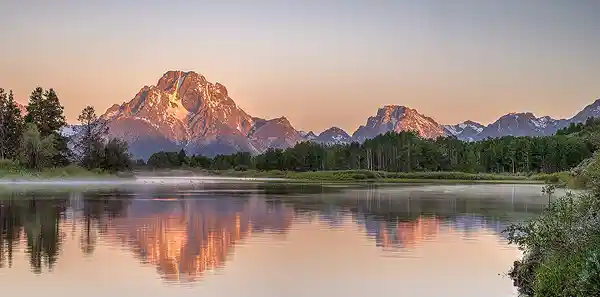 Sunrise Reflection of Mount Moran - Oxbow Bend Panoramic sunrise photo of Mount Moran reflecting in the Snake River at Oxbow Bend, with warm morning light and a peach-tinted sky. Taken by Mark Leatherman.