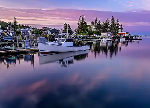 Port Clyde Morning Magic Sunrise in Port Clyde, Maine with a lobster boat at the dock, a red harbor building, and a pink-purple sky over a quiet coastal scene. Image by Rick Thompson.