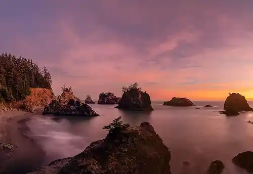 Sunset on the Oregon Coast Long exposure sunset at Secret Beach, Oregon Coast, with misty waves, pink clouds, and tree-topped sea stacks silhouetted against a colorful sky. Image by Bob Hart.
