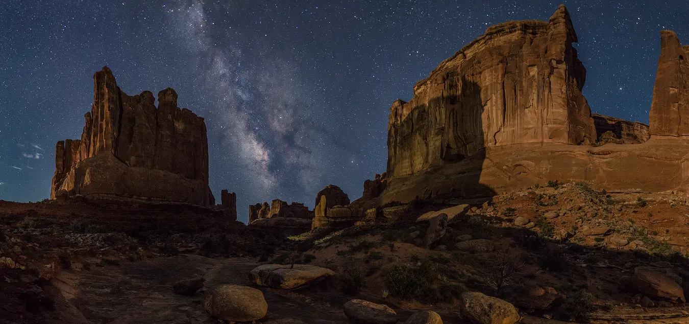 In this night panorama, the Milky Way is almost perpendicular in the sky and framed between two foreground buttes. Several more buttes are also framed and in the background. Because of their distance in the scene these buttes are much smaller and shorter and are positioned right below the galactic core. The foreground shows several car sized boulders spread out in a wide, dry creek bed. What make this image particularly beautiful is that a dim moon has risen, out of view to the left, casting interesting side lighting on the buttes and bluing and lightening the sky just enough to contrast nicely with the warm tones of the desert landscape.