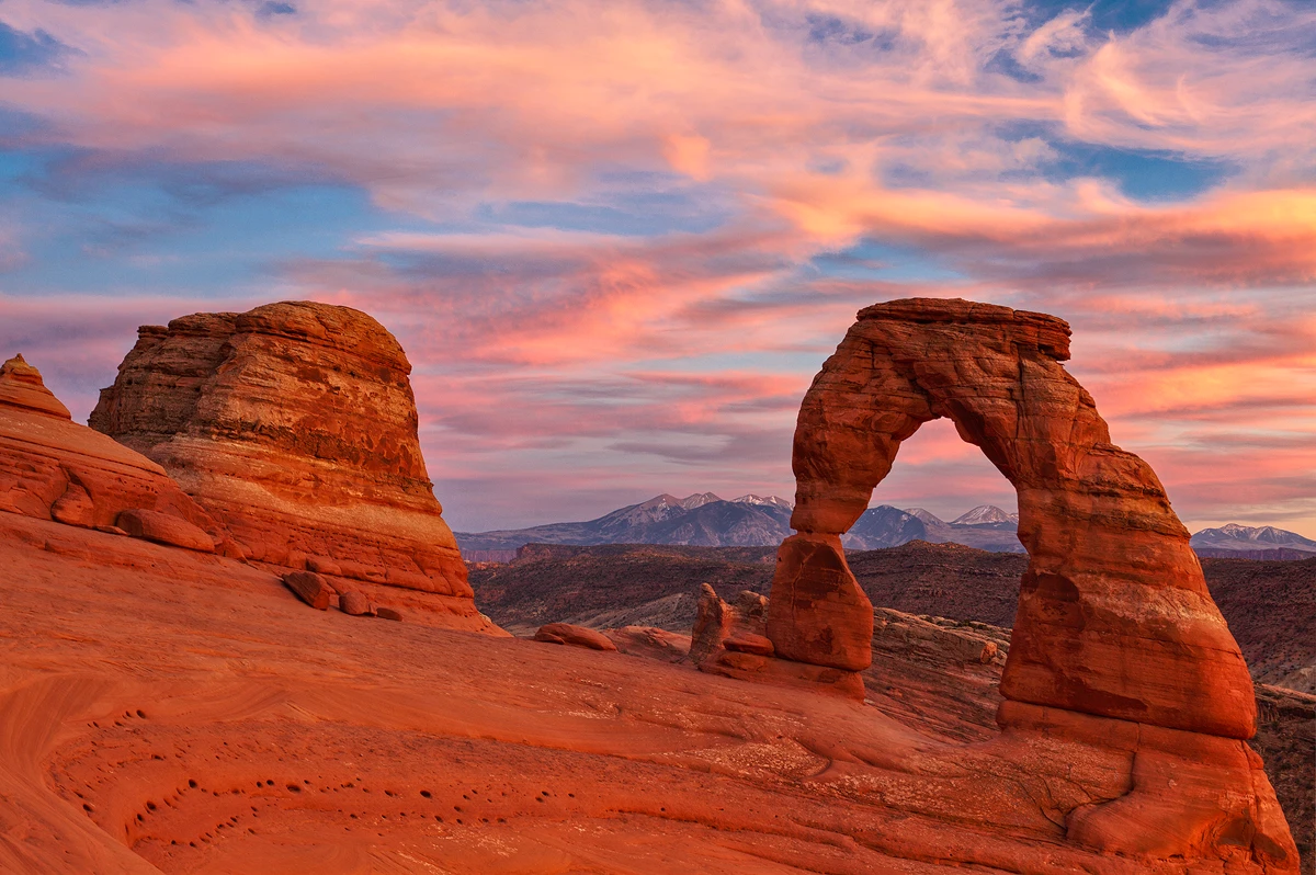 The view is to the east, the sun is setting behind the camera. The sky is the most overwhelming part of this image, it is full of lenticular clouds reflecting a range of pinks and yellows that contrast marvelously with the blue gaps between them. The red rocks of Delicate Arch and it's natural amphitheater are in the bottom half of the image and here they glow orange with the warm reflected light from the sky. This is the classic composition which shows the distant La Sal Mountains with early winter snow positioned left and right behind the arch and being visible inside the arch.