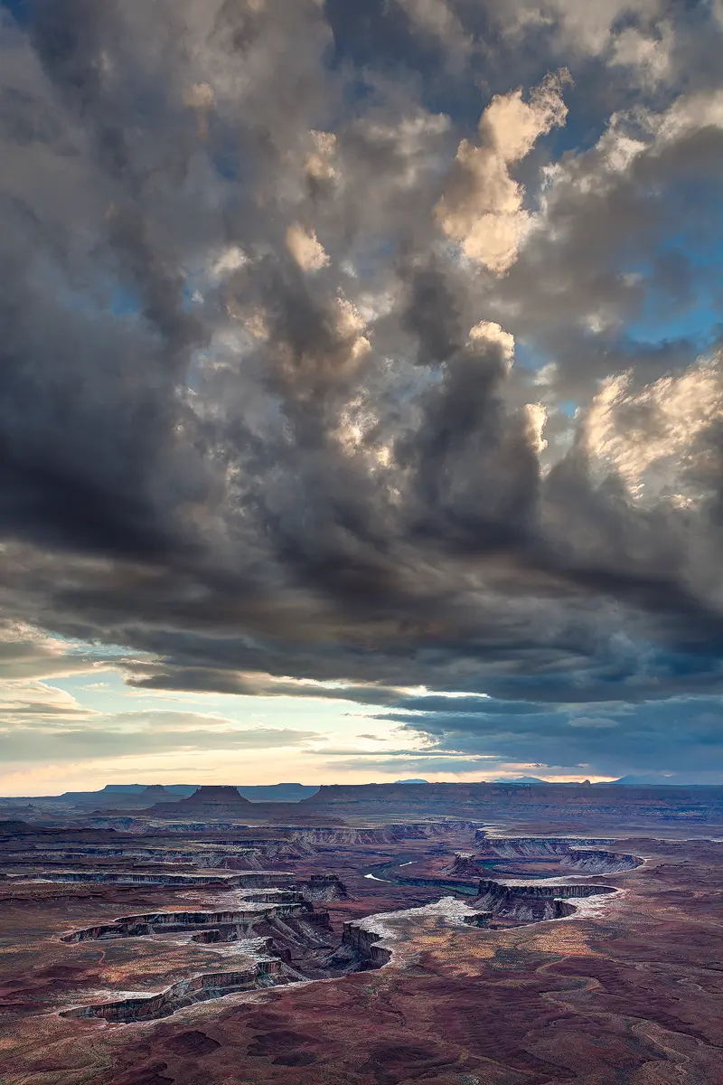 The top three quarters of this image is filled with stormy, dramatic clouds that remind one of an upturned hand with the wrist out of the frame to the right and pointed toward the sun. Below, in shade, and at distance, one sees a white rimmed canyon and hints of the Green River as it flows through Canyonlands National Park.
