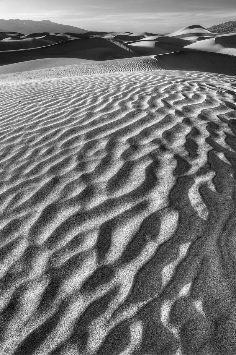 This is a wide angle, black and white, vertical image taken in the Mesquite Flat Sand Dunes of Death Valley National Park. The sun is at a low angle making shadows in the valleys of the sand ripples. Three quarters of this image shows the pronounced ripple pattern starting in bottom of the frame and it leads up to the top quarter which has a mass of dunes, then a sliver of mountains with a bit of cloudy sky at the top of the image.