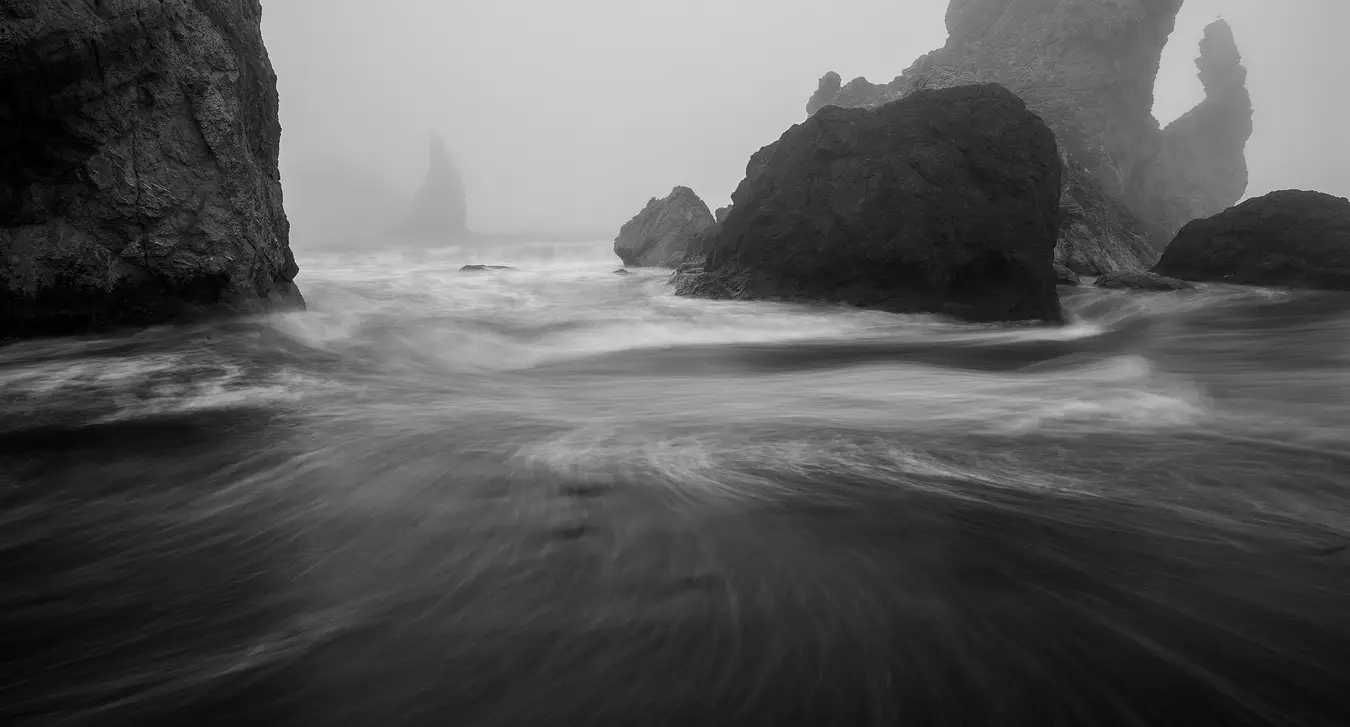 It's a stormy, foggy day on this rocky section of the Oregon coast. This black and white image shows nearby, large rocks that extend up from the water and out of the top of the frame. Positioned a bit left of center there is a gap between these rocks, there, in the fog, one can see the outline of a claw like rock spire. Behind it and further back, more obscured by fog, is a hint of another large rock formation. In the foreground, in the bottom half of the image, a longer exposure causes the surface froth on a retreating wave to record as streaks and swirl patterns that please the eye and lead it up from the bottom to the rocks and distant spire.