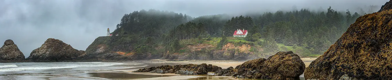 Stormy day panorama of the Heceta Head Lighthouse and lighthouse keepers home as viewed from Heceta Beach. This image shows a line of barnacle covered rocks sweeping in from the right side of the image and diminishing in size as they move away from the camera across the beach toward the ocean. About midway through the image the rocks dwindle away, then on the left, one sees the ocean lapping on a sandy beach. Above the beach are forested Oregon coast hills whose tops are shrouded in clouds. On the left side of the image, on a point above the ocean, sits the Heceta Head Lighthouse. Just to the right of the center of the image, in a forest clearing, is the light keepers house which is painted white and has a red roof.