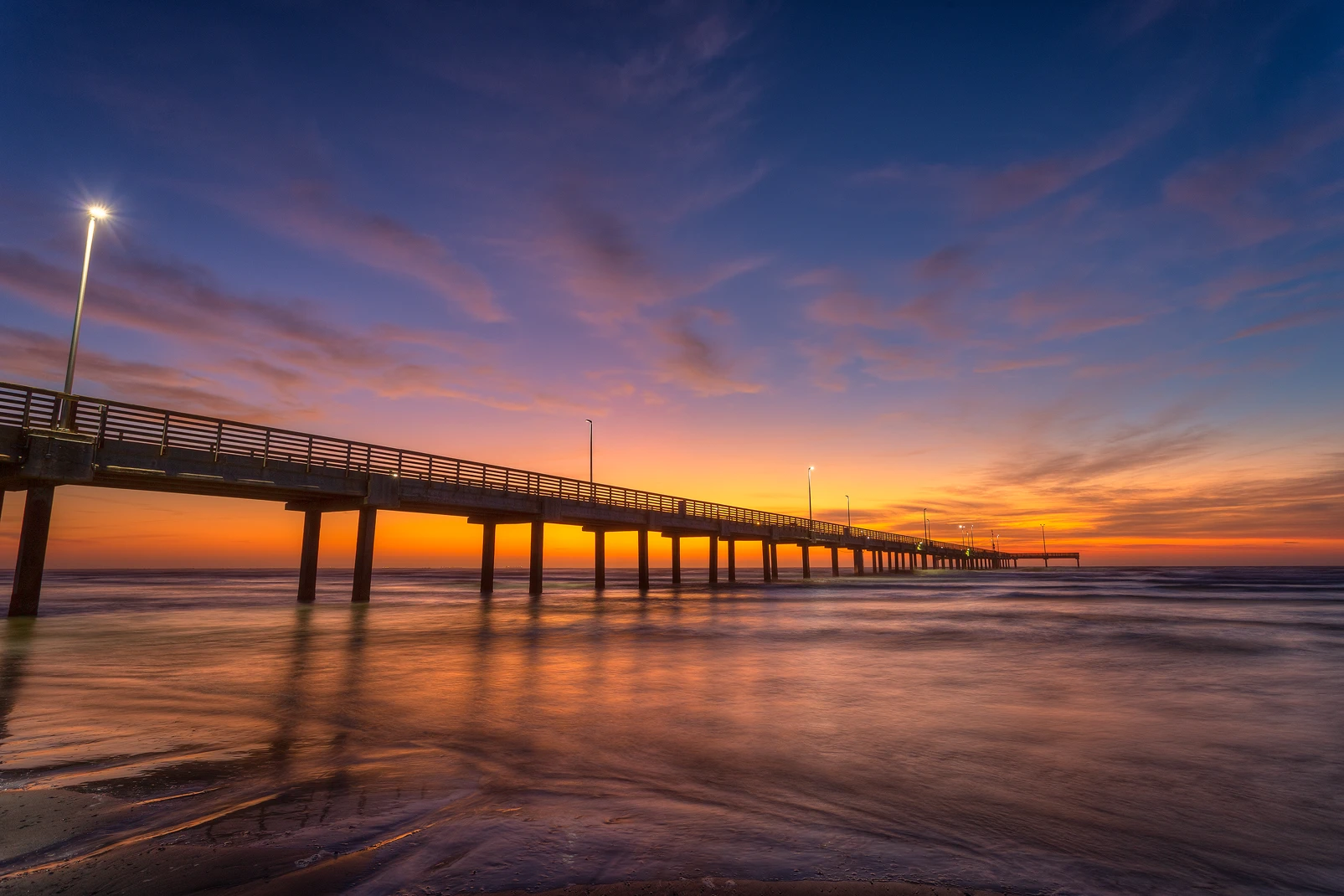 Horace Caldwell Pier sunrise photograph, Port Aransas, Texas: vibrant orange and blue sky, pier extending into Gulf of Mexico. Photo by Andy Cook.