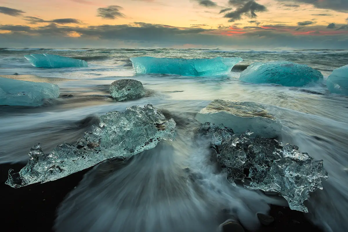 This image of a black sand beach faces sunrise and warm colors are filling the partly cloudy sky in the upper fifth of the frame. In the rest of the image the beach of black sand is mostly covered by an incoming wave which has to make it's way around large chunks of aqua colored glacier ice which are the size of footstools and bathtubs.