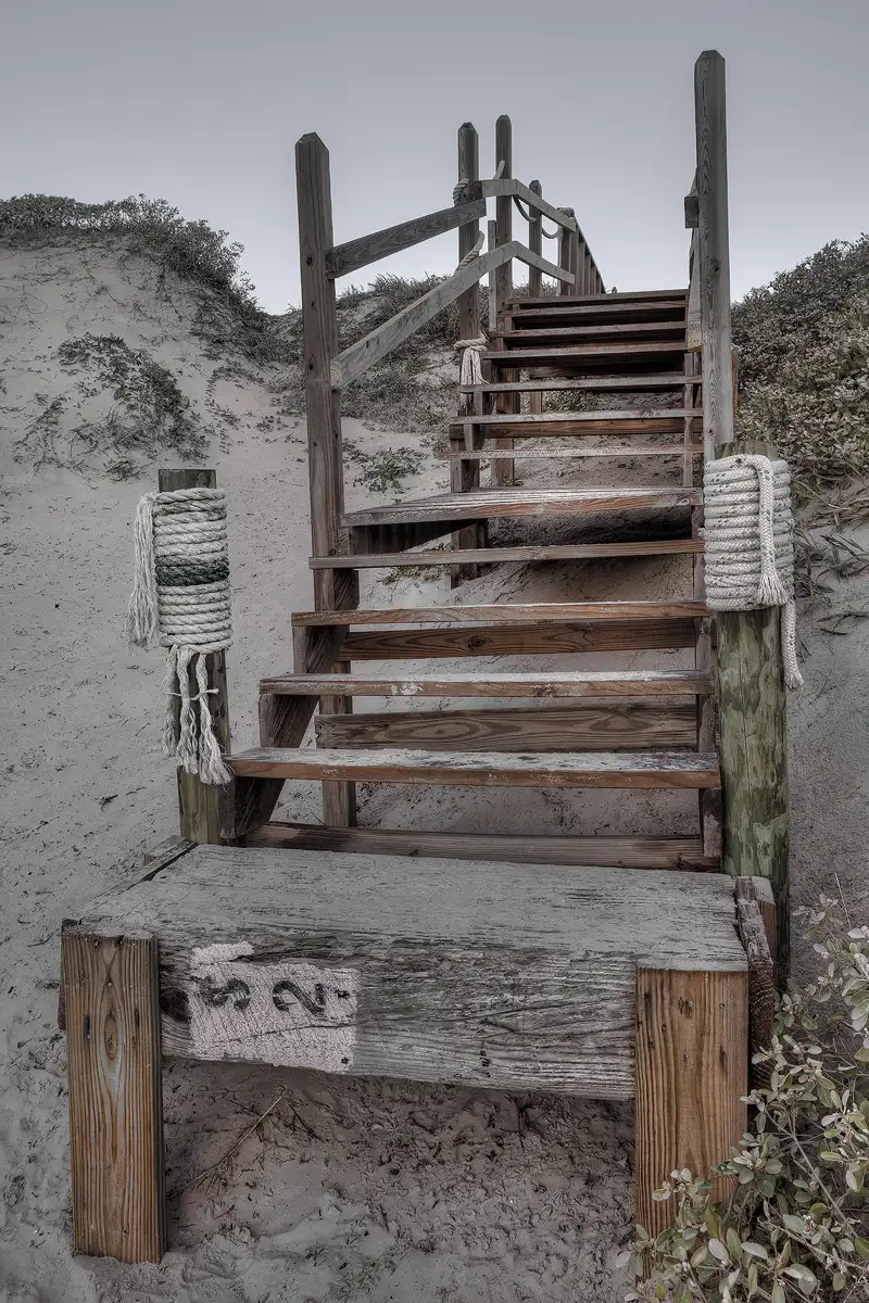 This is a rather simple vertical image. The frame is filled with a set of weathered, wooden stairs leading to the crest of a rolling, vegetation topped dune. The cloud free sky occupies the top one fifth of the image and the stair hand rails project into it. The way the colors have been processed make this a more interesting image. The colors are quite subdued, as if a color image and black and white image have merged.