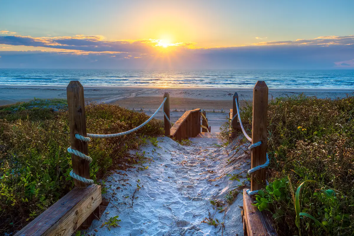 This sunrise image looks out over the Gulf of Mexico from the top of tall dunes. In the foreground, the lower half of the image, there is a sandy path emerging from the bottom center and heading up in the frame and back in the composition. Each side of the path has a big rope suspended from wooden posts and behind is thick, low vegetation. Near the center of the image the path disappears as it transitions to stairs and descends out of sight. Further back and higher in the frame is a sandy beach then the sea. Above, and left of center, the sun is just breaking through clouds and is giving a warm, yellow glow to the sky around it.