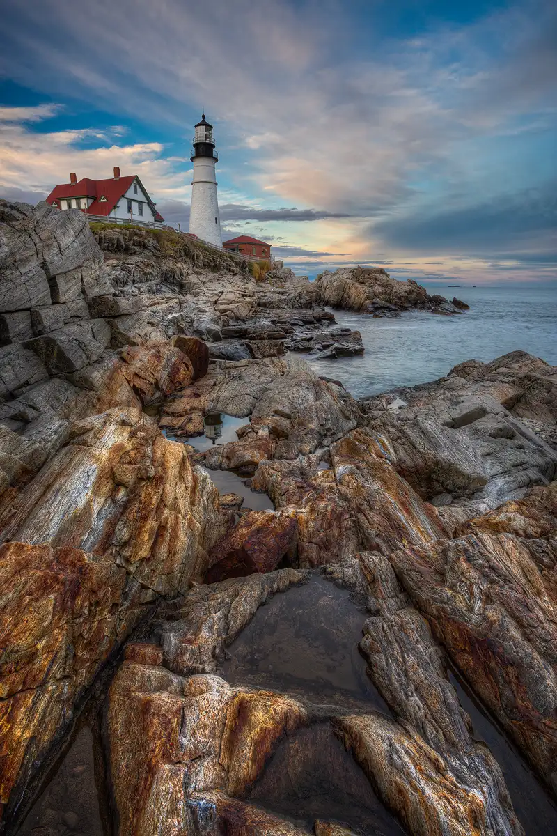 The sun is no longer shining in this vertical scene. It has set, but the striated clouds in the sky, in the upper one third of the frame, are still lit with warm hues. Projecting into this sky, left of the middle of the frame, is the tall, white tower of the Portland Head Lighthouse, capped by it's black top. This tower sits on a cliff above the ocean, the red roofed keepers house, partly obscured by the cliff, is to it's left. To the right there is a small red brick building and then rocks and the ocean. The bottom two thirds of the image shows the rocky coast leading back to orange and white striated rocks and water puddles in the foreground. The striations in these rocks point toward the lighthouse.
