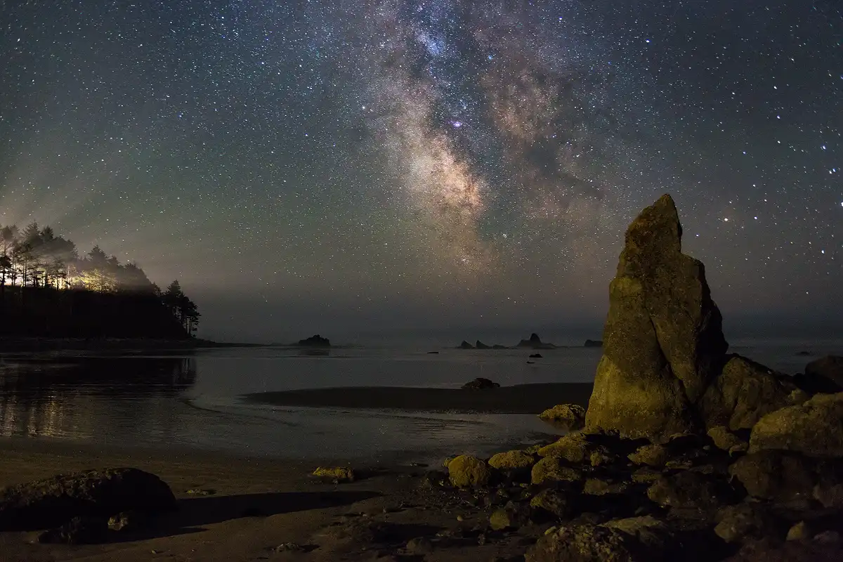 It is late at night on the Washington coast in Olympic National Park. The galactic core of the Milky Way is visible at an eleven o'clock angle in the upper three fifths of the center of the image. Framing it in the sky, on the right, is a rock spire emanating from the boulder strewn, sandy beach. Moving left, under the Milky Way, there are several rock spires out at sea, in the distance. Framing the Milky Way on the left is a forested hillside which is particularly interesting because a bright light in this forest is causing light beams to streak out through gaps in the tree branches and shine toward the Milky Way.