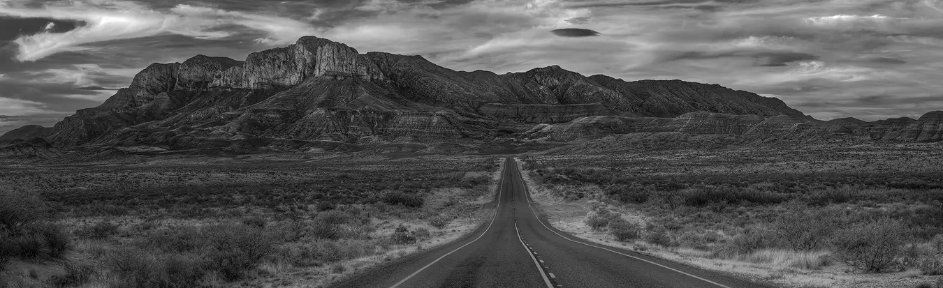 An empty two lane highway enters right of center in the bottom of this black and white panorama. The terrain of the low bush desert has it declining away from the camera for a short way before starting a gradual incline. The road continues straight and it narrows as it moves further away. In the distance, where the width of the road is very small and about half way up the frame, a dramatic looking mountain range arises. Above it beautiful cloud patterns play in the sky.