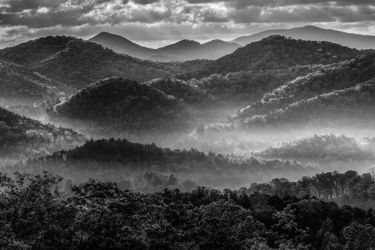Long lens, black and white photograph of layers of back lit, rounded hills leading up to taller ones in the background. The upper one sixth of the image shows a mostly cloudy sky, but direct sunlight is getting through many gaps causing the top edge of some hills to glow with a rim light creating distinct boundaries. It's still early in the morning and a misty fog flows between many of the hills and where sunlight shines, it glows brighter.