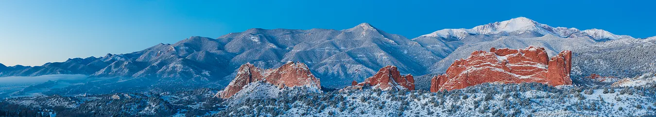 It's before sunrise on a winter morning and still in the blue hour, bluish light is cast over the scene. This panorama taken from distance with a long lens has two main features. The foreground, in the lower section, reveals a row, left to right, of three dramatic red sandstone rock formations in Garden of the Gods. The background, higher in the image, above the Garden of the Gods rock formations, shows the front range mountains from Cheyenne Mountain on the left to Pikes Peak on the right side of the image. What make this image particularly appealing is that a layer of fresh snow coats everything.