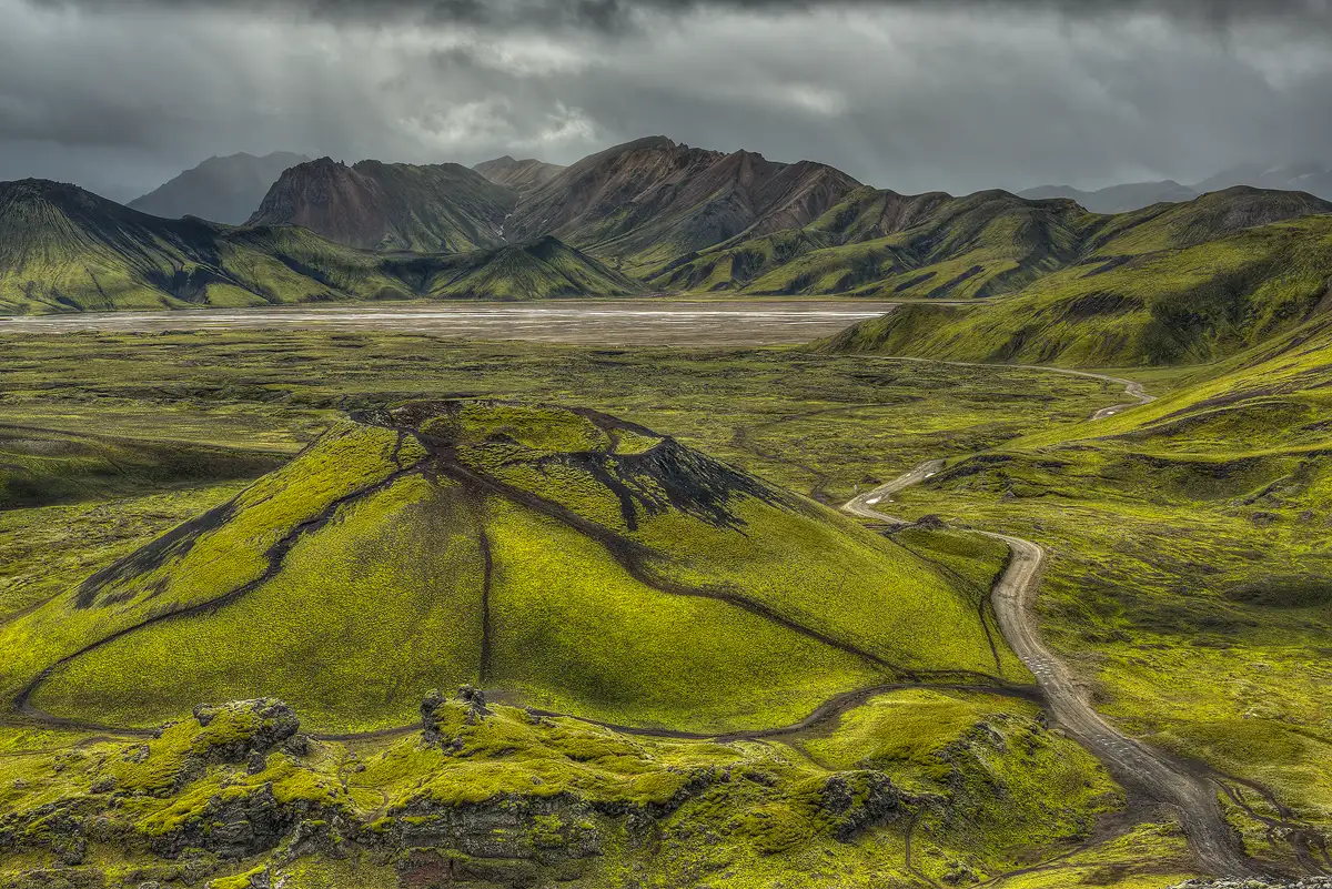 Iceland has many areas where it receives enough moisture that in summer a short, green, mossy collection of plants covers the landscape, but it takes a while to grow and often it's missing on some steeper hillsides and rocky outcrops. Well, that's the feel of the bottom four fifths of this image taken from a high vantage point looking down and out. In the foreground, occupying the left two thirds of the frame is a large, conical hill with three foot paths climbing it from different angles. Near the bottom right corner of the frame a dirt road enters and leads the eye past the right side of the conical hill to a flat area behind it and on back in the image where one finds gnarled looking storm clouds scraping across distant mountains.