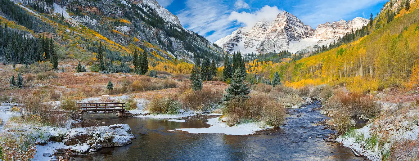 Steep, nearby mountainsides, left and right, create a u shape valley in the upper half of this autumn panorama. This valley is right of center in the image and it frames the spectacular Maroon Bells in a partly cloudy, morning sky. Maroon Bells is a set of pyramid shaped mountains that are very close to each other with the right one being a bit nearer. The valley has a stream flowing toward the viewer then it curves left, under a foot bridge, then out the left side of the image. This valley also has meadows and stands of spruce and golden aspen trees, but what really makes this image special is the fresh layer of snow that had fallen during the previous night.