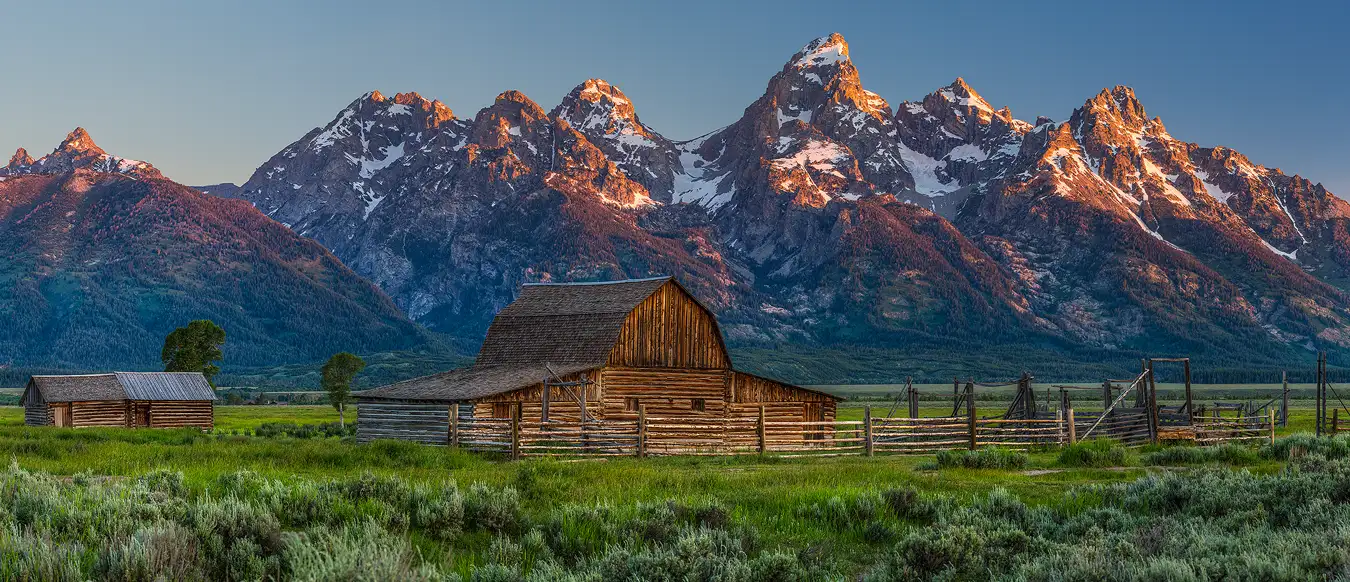 A rustic, western barn with a corral faces the viewer. It sits in a green, sage brush field. Behind it and rising high above it are the dramatic Grand Teton Mountains, their peaks capped by snow and receiving the days first warmly hued rays of sunlight.