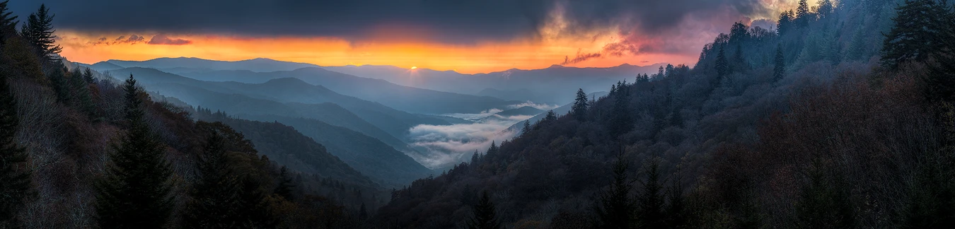 This dawn panorama is from a high elevation facing east and down a v shaped valley with layers of blue hued, mountain ridges repeating the v shape into the distance. Fog lingers at distance in the valley. Just left of the middle of the frame and about three quarters of the way up a sliver of the sun's disc is just cresting a distant mountain ridge. A band in the sky, immediately above the distant ridge, is filled with orange and pink color. Above the color band the sky is filled with dark, ominous clouds.