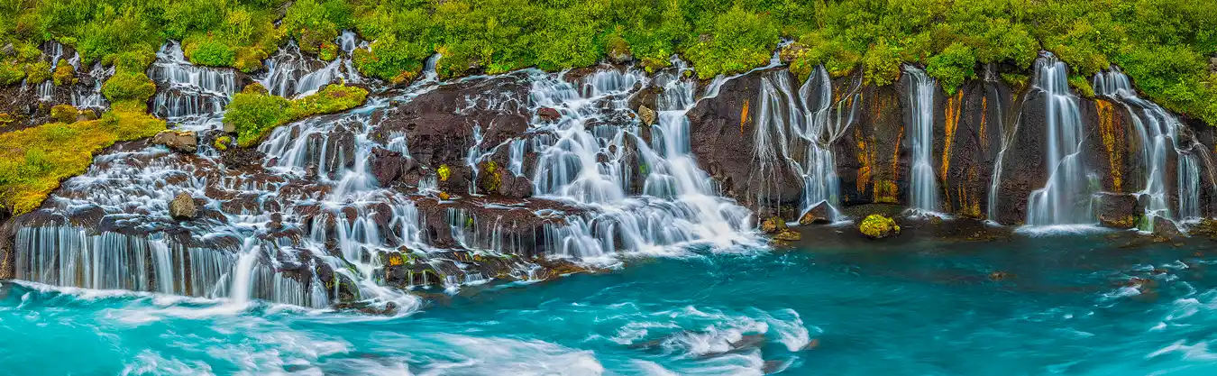 This is a long lens, panorama of a waterfall that flow out from under dense vegetation onto a rocky slope breaking into many streams and cascades before splashing into a blue-green river.