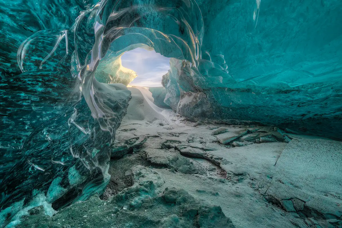 This image was taken inside of an ice cave carved by flowing water. Most of the image shows the sculpted, blue glassy walls and ceiling of the cave, but just off center, above and left of the middle, is a small opening and one sees sunlight shining on the left edge of this opening and a partly cloudy sky visible high above. Snow has drifted in and is piled on the floor below the opening.