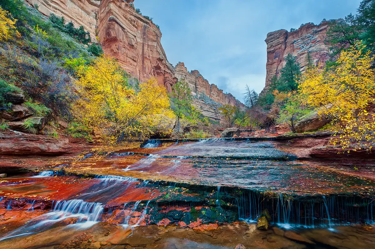 A wide shelf of red rock is across the bottom half of the image and it slopes down toward the viewer. A stream flows over this sloping expanse of rock emerging in the center of the frame and fanning out to fill the width of the frame at the bottom. The stream has many little cascades as it flows over layers in the shelf. On each side of the stream are vibrant autumn colors and above them, in the background, are sheer canyon cliffs that merge right of center in the frame forming a v shape with the stormy sky.