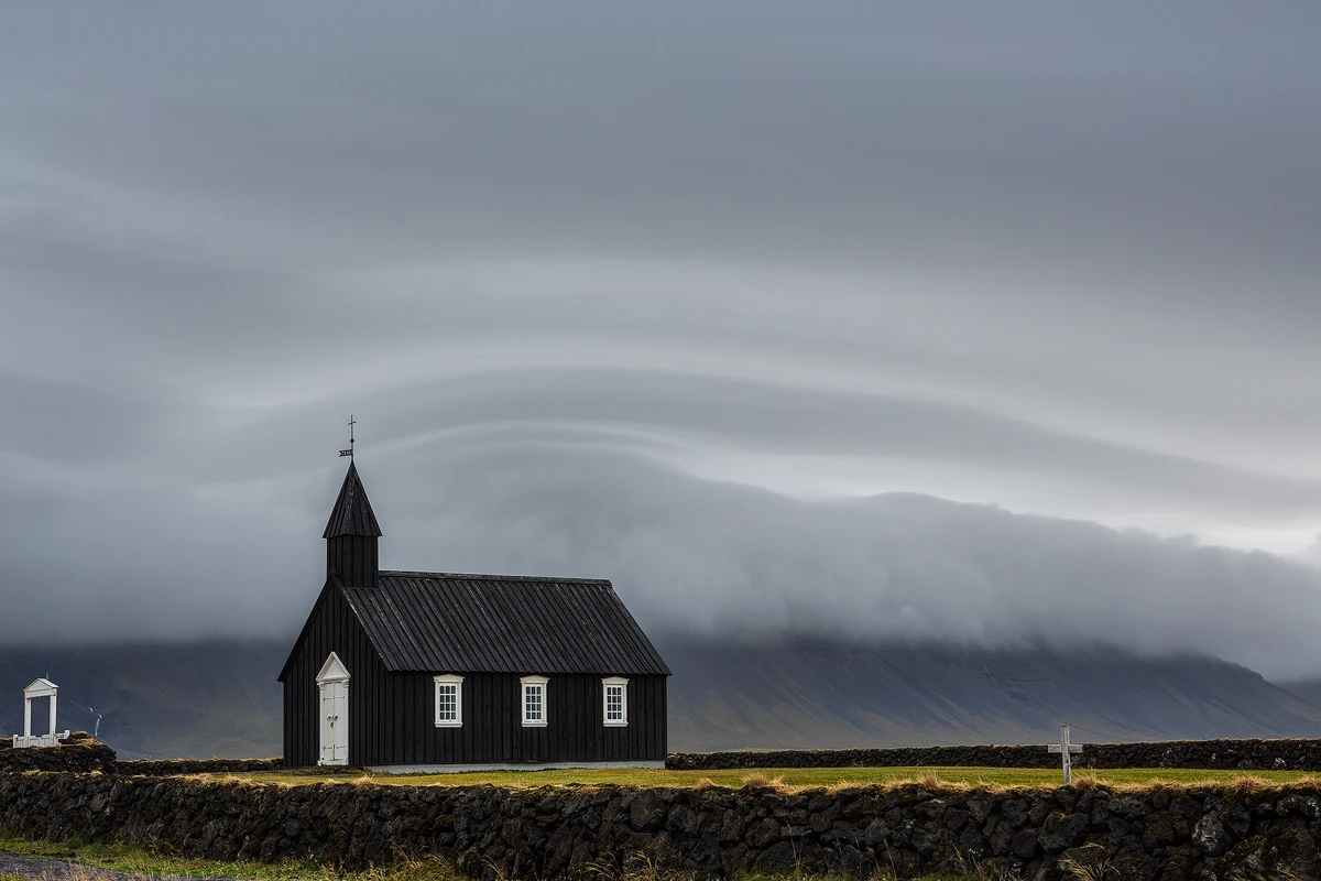 Photograph of the black church of Budir, Iceland. It is positioned in the lower, left power point of the image and faces out of the frame. There is a long, black stone wall below, in front of, the church. Behind and above the church is a curving cloud pattern hanging low over the tops of distant mountains and occupying the majority of the frame.