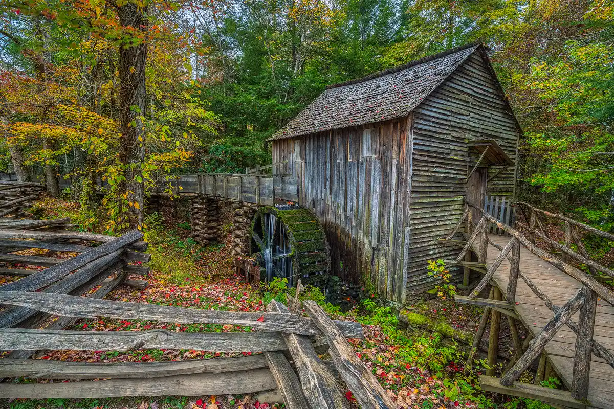 A zigzag fence cuts across the foreground on the left side of this image, in front of the grist mill. In the bottom, right corner an elevated, wooden ramp enters the frame and leads to the Cable Grist Mill that fills the area right of center in the frame. On the side of the mill, behind the zigzag fence, is a large, stationary, moss covered water wheel with water flowing down, through and around, the spokes and spindle. Autumn colors are on the ground and in the forest that surrounds the mill.