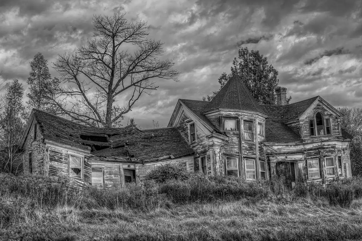This creepy, black and white image shows a classic looking haunted house along U.S. Route One in rural Maine. A summer's worth of long, overgrown grass fills the front yard, out of control shrubs push up against the walls and under the windows, some of which still have glass. The sky above the house is filled with brooding clouds and a leafless tree skeleton. The once prosperous house, with a rounded turret in front, has terminal foundation problems causing the home to lean and sway in every direction. The exterior paint, once white, is now mostly missing, replaced by weathered wood.