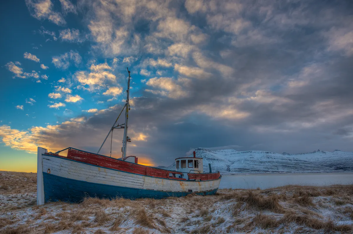 This winter image, the near the shore of an eastern fjord in Iceland, is comprised of long brown grass dusted with snow. On this grassy shore sits an old, wooden hull fishing vessel. Long ago the hull was colorfully painted with three horizontal stripes, a red one on top, white in the middle and blue at the bottom. Across the fjord snowy mountains rise to the morning sky filled with beautiful clouds receiving early light.