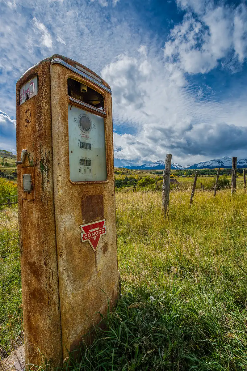 Photo of an old Conoco gas pump on Last Dollar Road near Telluride, Colorado. The image was taken from mid pump level and the pump fills most of the left side of the frame. The pump is set in an overgrown meadow beside a wire and wood post fence and it's very old, mostly rusty with flaked white paint. The background shows snow capped Colorado mountains and a beautiful, partly cloudy sky.