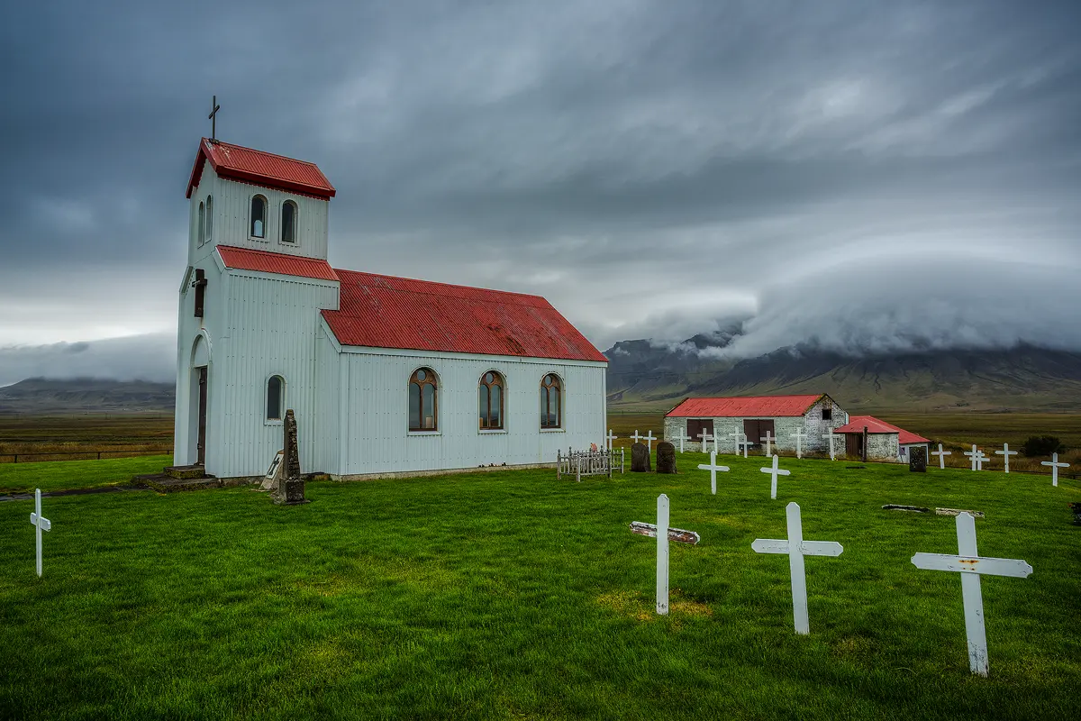 On a grassy plain in front of distant mountains being raked by stormy clouds sits Miklaholt Church. It is a red roofed, white church with a red roofed, white outbuilding far behind it and an open, flat, grassy graveyard with white, wooden crosses on one side and immediately behind the church.