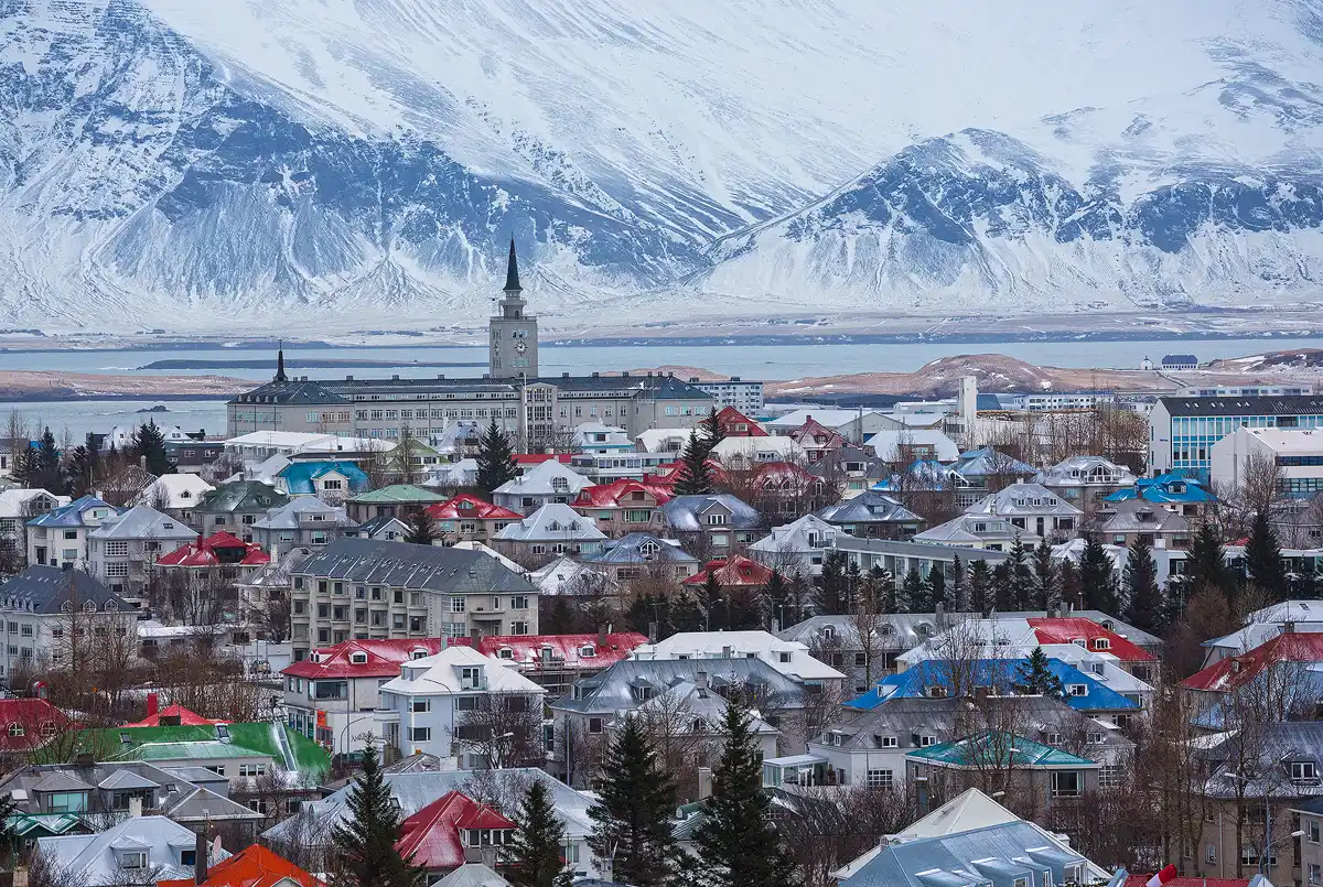 This long lens image, from a higher elevation looking down, shows the colorful rooftops of Reykjavik in the bottom half of the frame then Faxafloi bay with distant mountains above it, these mountains exit the top of the frame. A recent snow lingers on the background mountains and in places on many of the colorful roofs.