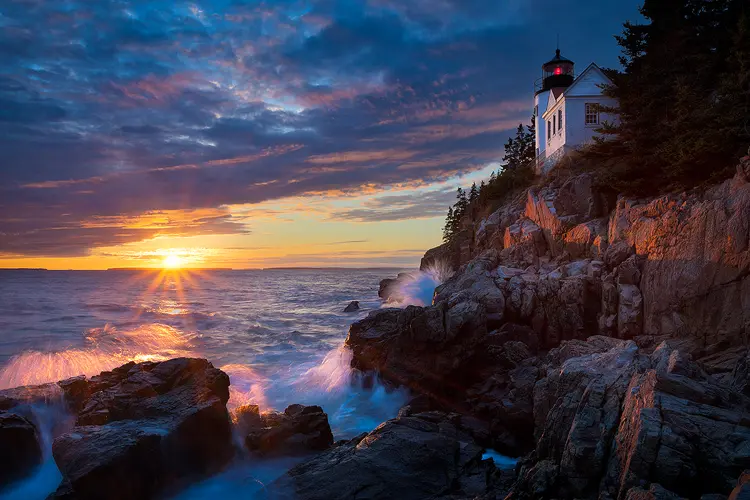 The sun is setting beneath beautiful clouds and over wavy water in the left side of the image. In the upper right, on a cliff above the water, the Bass Harbor Head Lighthouse receives the last few sun rays of the day. Below, waves break on rocks at the foot of the cliff.