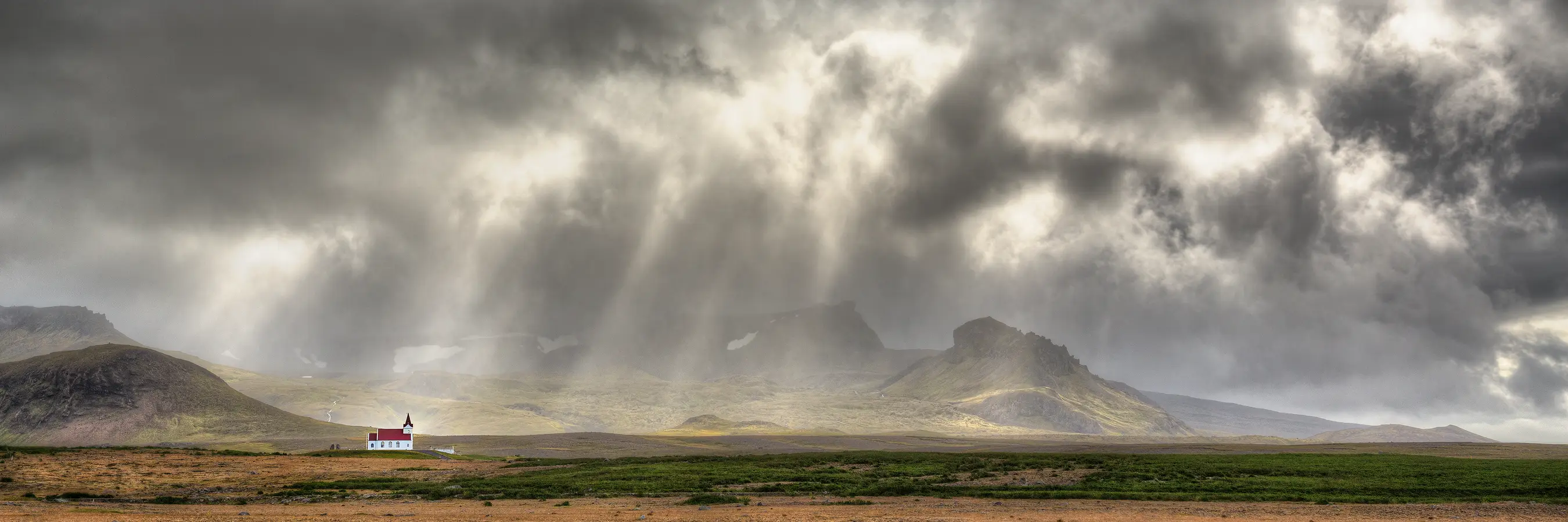 Storm clouds still roil, but gaps open and shafts of sunlight streak through the turbulent sky. Below, in the bottom third of this panorama is a vast plain in front with background mountains disappearing into the clouds. On the left side of the image, a white church with a red roof sits at distance, facing right, to the west, as is the custom in Iceland. It's alone, on the otherwise empty plain.