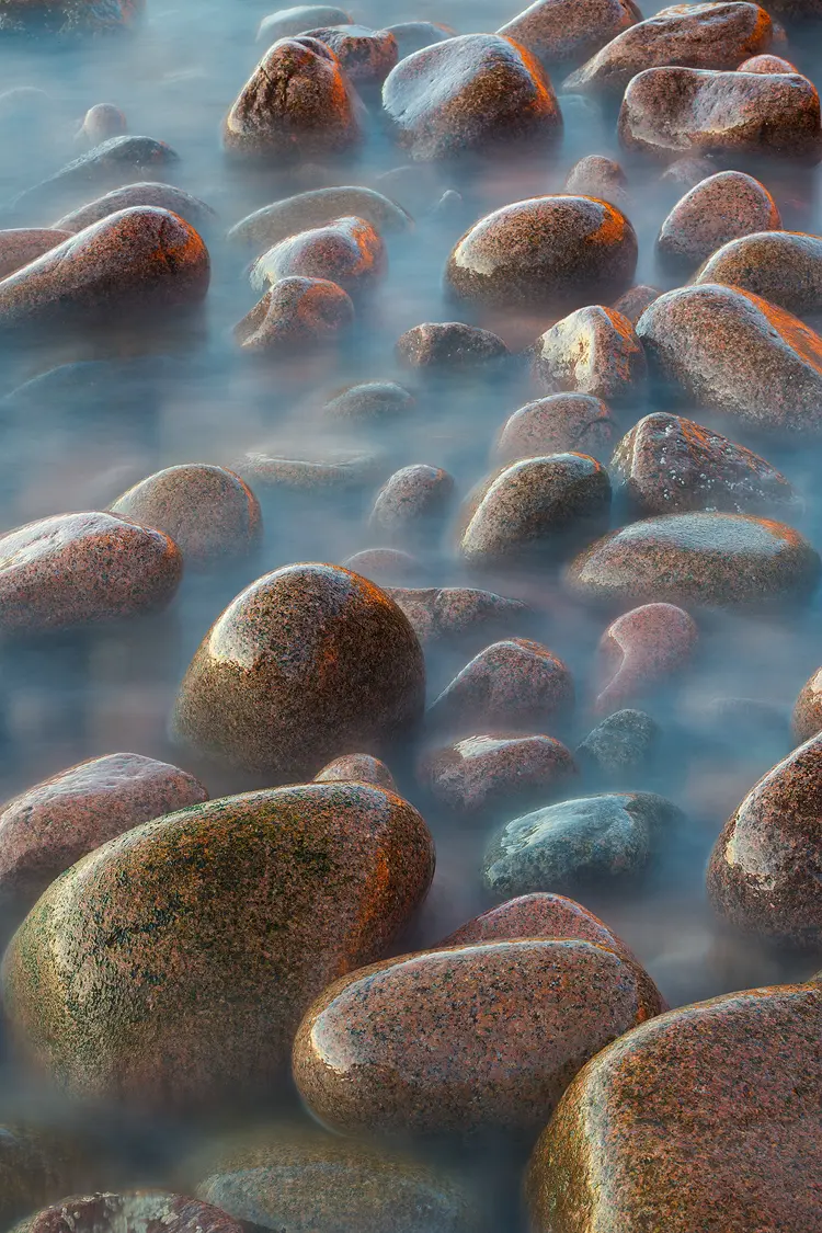 Photography location; Long lens, vertical photo of smooth boulders on a beach in Acadia National Park, Maine. The boulders are getting washed by waves and a long exposure time has caused the waves to record as an ethereal looking fog surrounding the wet, shiny boulders.