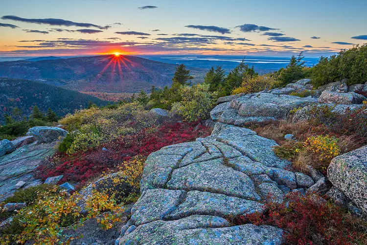 Workshop location; Autumn photo of sunset from Cadillac Mountain in Acadia National Park, ME. The bottom half of this image shows mostly pink and black granite with green and blue lichen covering much of it. The granite has various shelves and large linear cracks. Interspersed in the granite are various red, yellow and green bushes. Above, in the background, are layers of mountains. The sun is radiating a red starburst as it sets in the summit of a mountain on the left side of the frame.