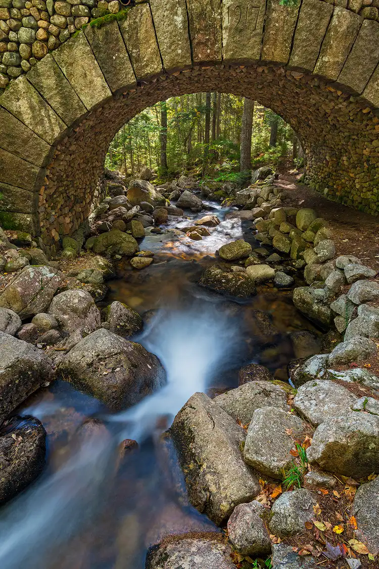 Workshop location; Vertical photo of the Cobblestone Bridge in Acadia National Park, Maine. The stream that flows under the arching bridge, covered in cobblestones, enters the bottom left of the frame flowing away from the camera and under the bridge. Framed by the bridge’s arch one sees a forest and the stream twisting out of sight.