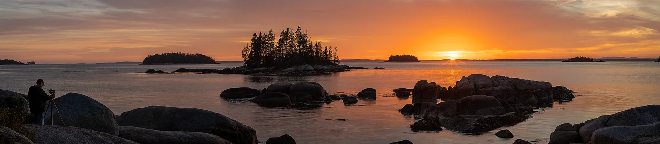 Workshop location; The sun is setting and the sky is filled with orange clouds in this panorama from the coast near Acadia National Park. In the foreground are many large, granite rocks surrounded by water reflecting the orange clouds. Looking across the water, in the middle ground, one sees many small, treed islands and they continue to be visible, but get smaller in the background.