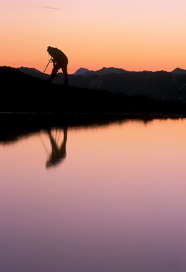 Silhouette, against a pre-dawn sky, of a photographer and his reflection in a mountain lake.