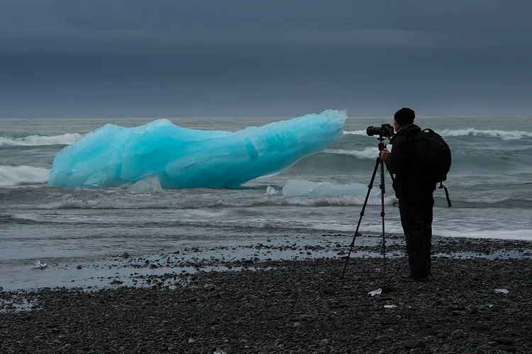 A photographer on a black stone beach in Iceland photographing blue chunks of glacial ice.