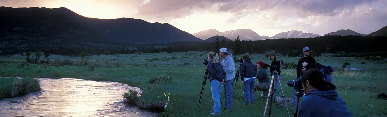 Panorama of a group of photographers in a field next to a stream photographing sunset.