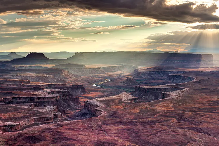 Workshop location; This image shows the view from Green River Overlook in Canyonlands National Park, UT. In the sky, broken clouds allow warm, light beams to shin down on the scene from the late day sun. The scene below shows a reddish rocky plain where eons of erosion have allowed the Green River to carve twisting canyons. The rim of the canyons have eroded down a little lower than the surrounding plain causing a white rock to be exposed and outline the rim of the canyons. Fading into the distance one sees layers of ridges and mesas.