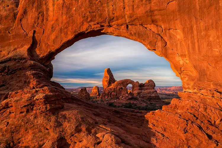 Workshop location; Sunrise light is causing the reddish rock that makes up the North Window in Arches National Park to glow orange. The entire frame is filled by the arch, rock exits every side of the image. The opening of the arch is centered in the frame and inside one sees Turret Arch and the landscape around and behind it as well as the cloud filled sky above the arch.