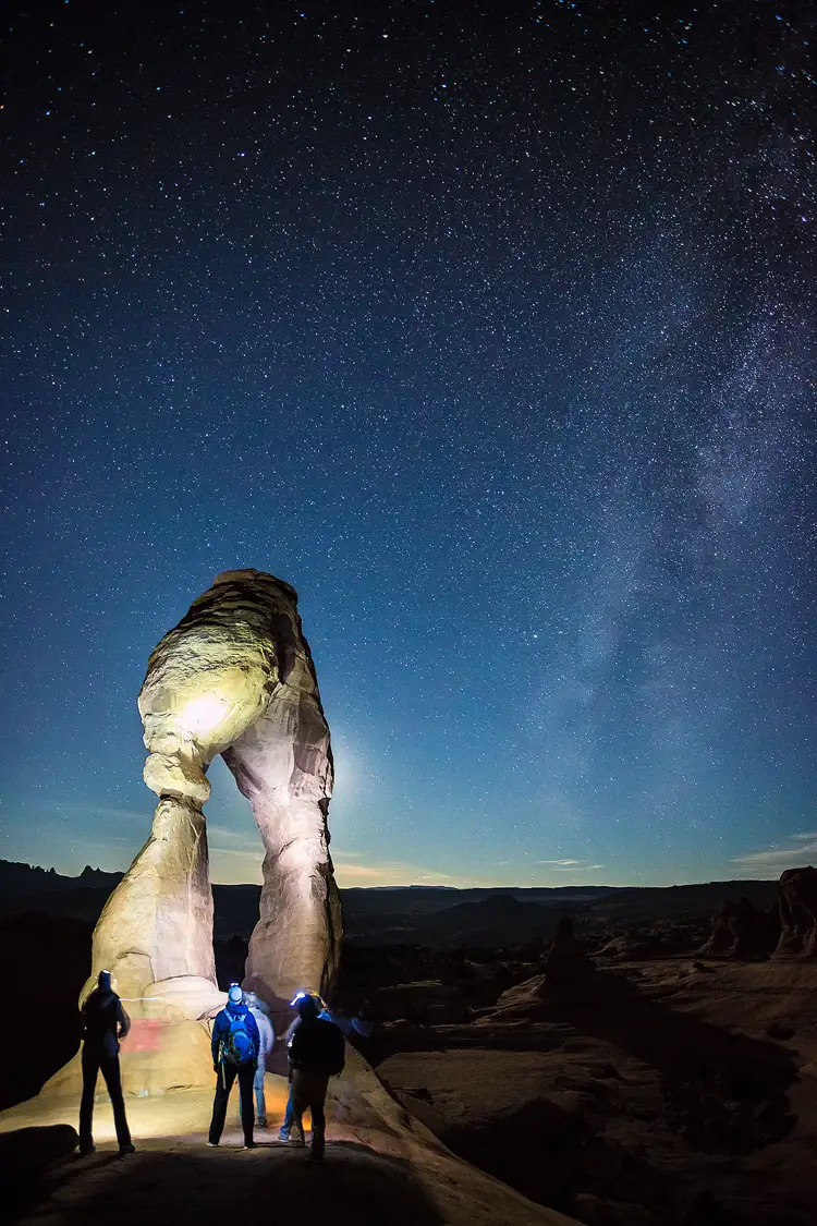Workshop location; A handful of hikers wearing packs and headlamps face away from the camera looking at Delicate Arch at night. Their headlamps light the arch and silhouette some of the hikers. The dark sky is still blue, but the stars and Milky Way are becoming visible.