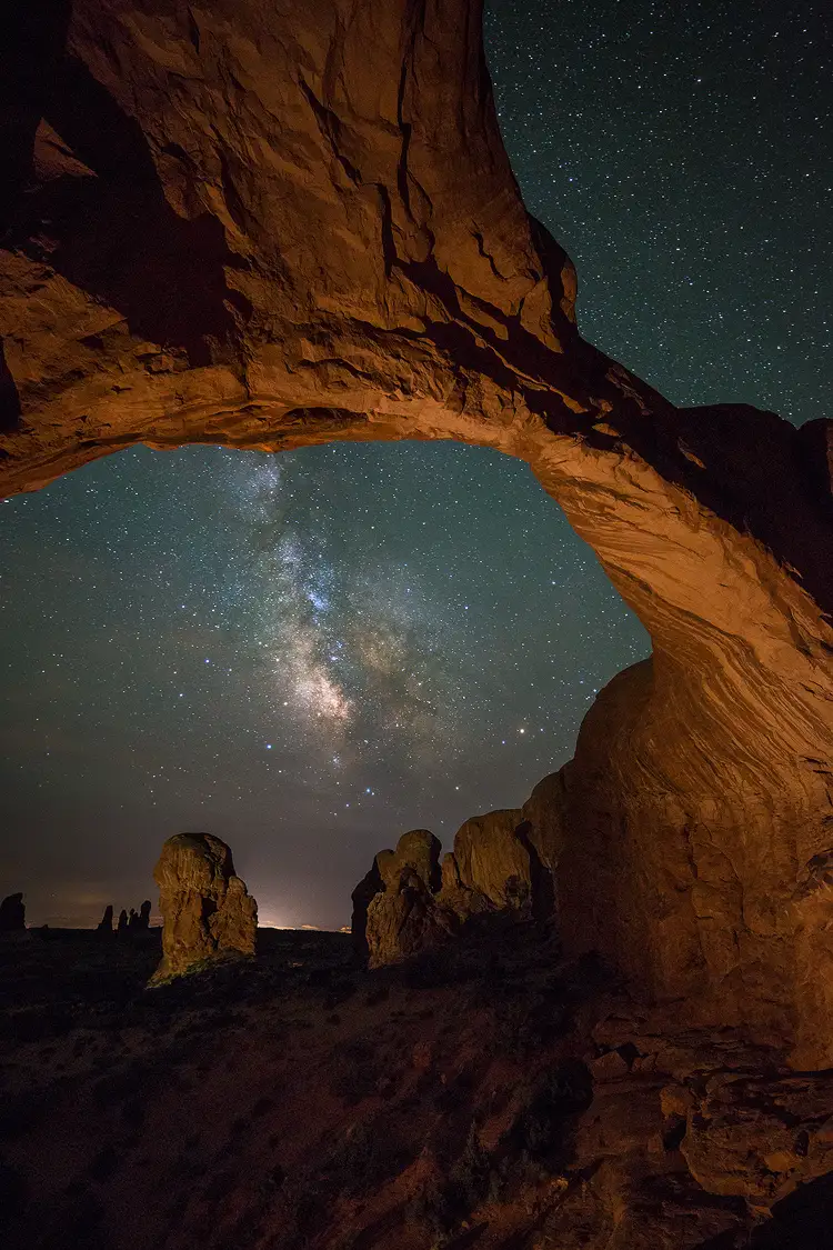 Photography location; In this vertical, night photo of Double Arch in Arches National Park, the camera is under the arch and pointed up. This brings the arch in from the upper left corner of the frame and down toward the bottom right, where it's anchored to a rock wall. In the large opening in the center of the frame, between the arch and the ground, sits the Milky Way.