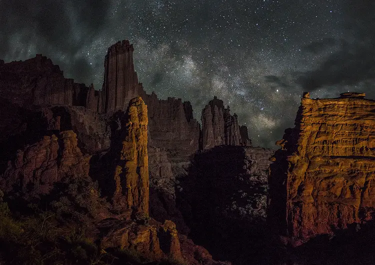 Photography location; At night Fisher Towers near Moab, UT stand tall and monolithic looking as side light brings out vertical, striated detail in the red hued pinnacles. In the sky, arching above the towers, is the Milky Way.