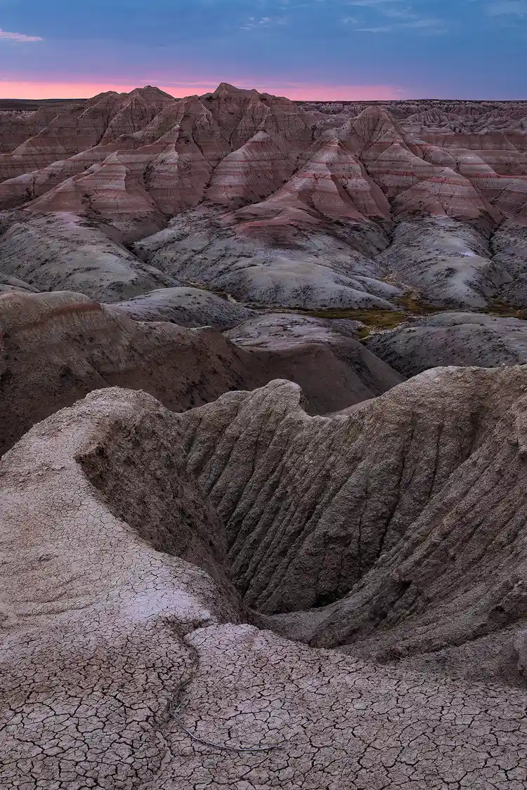 Workshop location; The bottom of this vertical image shows mud cracked soil on top of a skinny ridge that juts out, down and away from the viewer into layers of eroded canyons of crumbly rock and across a small valley to a rugged hillside of alternating layers of red and light gray rock.