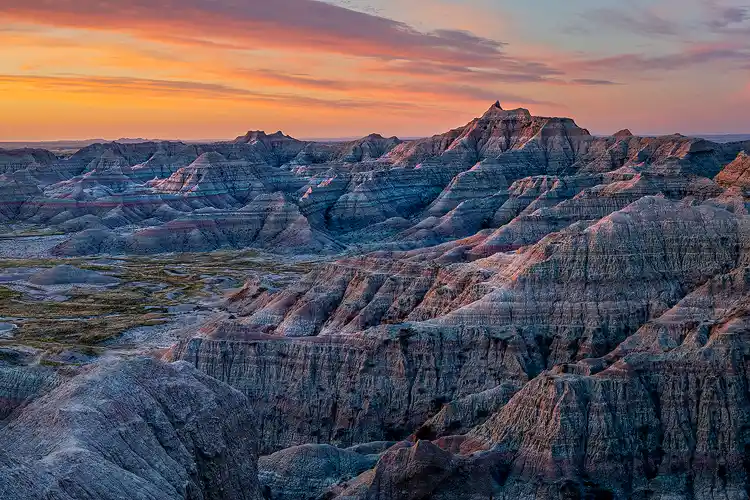 Workshop location; Streaks of yellow, orange and blue fill the sky as clouds collect the days first light in the top third of this Badlands National Park image. The bottom of the image has steep ravines and curving ridges which bring the eye in, and up, from the bottom left corner to the distant jagged, peaks which fill the middle of the frame and are beginning to glow orange with side light.